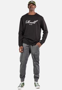 Schwarzer Sweatshirt mit "Reell"-Logo, graue Cargo-Hose, grüne Kappe und grüne/weiße Sneaker. Lässiger Schnitt mit gerippten Bündchen.