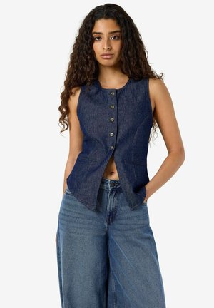 Femme aux longs cheveux bouclés portant un gilet en denim foncé sans manches boutonné et un jean bleu à jambes larges avec les mains dans les poches.