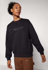 Felpa nera con lettering "tresor" in tonalità, colletto rotondo e polsini a coste. Indossata con pantaloni marroni dotati di tasche laterali e dettagli metallici.