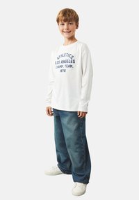 Biała koszula z długim rękawem z granatowym napisem "ATHLETICS LOS ANGELES CHAMP. TEAM. 1978", w zestawieniu z luźnymi niebieskimi dżinsami i białymi sneakersami.