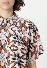 Floral button-up shirt featuring a cream background with red and navy patterns, short sleeves, and a pointed collar. Smooth fabric texture.