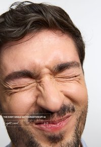 Nærbilde av en persons ansikt med sammenknyttede øyne og et svakt smil. Huden er lys, med ansiktshår, mot en enkel hvit bakgrunn.