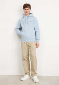 Light blue hoodie with a front pocket, drawstrings, and ribbed cuffs paired with beige pants and white sneakers. Casual cotton blend fabric.