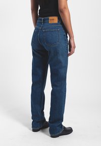 Blue denim jeans with a straight cut, featuring a leather patch on the back, dual rear pockets, and contrasting stitching along the seams.