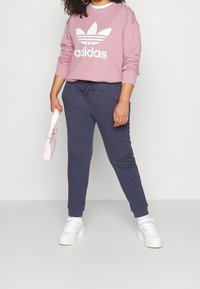 Różowa bluza z białym logo Adidas, w zestawie z granatowymi dresami i białymi sneakersami. Trzyma małą różową torbę. Kręcone włosy.