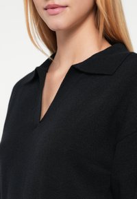 Pulover negru tricotat cu decolteu în V și guler plat, cu o textură netedă și o siluetă mulată.