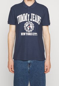 Mörkblå bomullspikétröja med krage, med en vit logografik som inkluderar "TOMMY JEANS" och "NEW YORK CITY". Korta ärmar.