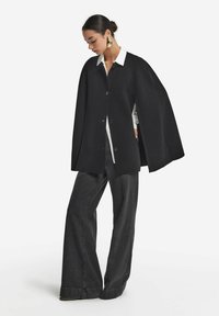 Femme portant une cape noire à boutons sur une chemise blanche et un jean large foncé, regardant vers le bas contre un fond uni.