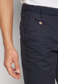 Gros plan d'une personne portant un pantalon bleu marine avec une ceinture tressée et une poche boutonnée, chemise blanche rentrée, main gauche détendue sur le côté.