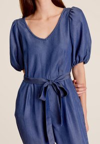 Denim jumpsuit met een diepe V-hals, pofmouwen en een strikceintuur in de taille. Heeft een zachte textuur en een donkerblauwe kleur.