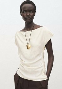 Top crème, à manches courtes et à coupe décontractée, présentant un tissu texturé. Un collier avec un pendentif en or pend autour du cou.