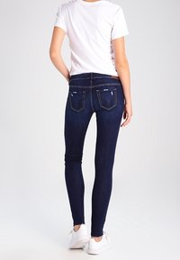 Jeans skinny azul oscuro con bolsillos traseros, ligeramente desgastados, y dobladillo a la altura del tobillo, combinados con una camiseta blanca sencilla y zapatillas.