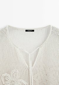 Biały top z siateczkowego dzianiny z dekoltem w kształcie litery V i haftowaną kwiatową ozdobą w pobliżu dołu, z metką Massimo Dutti wszytą wewnątrz kołnierzyka.