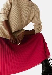 Beiger, gerippter Pullover mit langen Ärmeln kombiniert mit einem plissierten pinken Rock. Schwarze Schnürstiefel vervollständigen das Outfit.