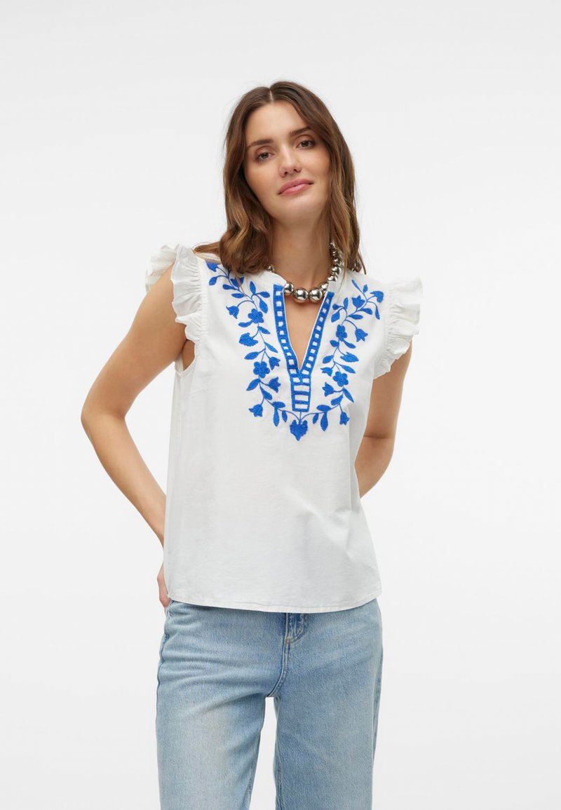 Haut en coton blanc avec broderies fleuries bleues et manches volantes, présentant un décolleté en V et des accents rayés bleus le long de l'encolure.