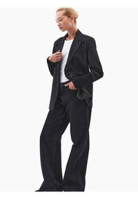 Nainen, jolla on päällään musta bleiseri ribbireunuksilla, valkoinen paita ja leveälahkeiset mustat farkut, seisoo yhdellä kädellä taskussa ja toinen käsi koskettaen bleiseriä.