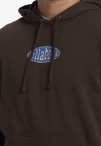 Sudadera marrón que presenta un logo azul y blanco en el pecho, con capucha ajustable y tejido texturizado. Corte regular con puños acanalados.