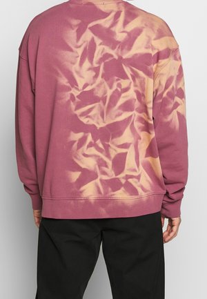 Person trägt einen langärmeligen Batik-Sweatshirt in Mauve und Beige mit schwarzer Hose, von hinten vor einem einfarbigen Hintergrund gezeigt.