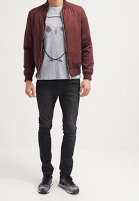 Chaqueta bomber burdeos sobre una camiseta gris con un gráfico de búho, combinada con jeans oscuros y zapatillas negras con estampado. Ajuste casual.