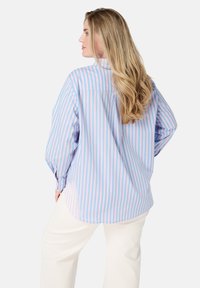 Chemise rayée bleu clair avec des rayures verticales roses, à manches longues, coupe ample et ourlet arrondi, associée à un pantalon crème.