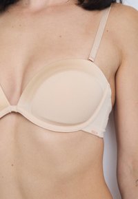 Sutiã acolchoado em bege com copas alisadoras, alças ajustáveis e um brilho sutil. Apresenta uma pequena etiqueta de logotipo perto da alça.