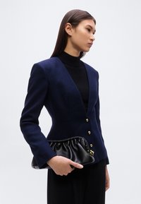 Μπλέ ναυτικό σακάκι ραμμένο, με υφή, χρυσά κουμπιά και εφαρμοστή μέση. Στο χέρι κρατιέται μια μαύρη τσάντα φάκελος με ρίγες.