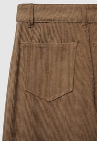 Pantaloni in velluto a coste marroni con texture a righe verticali. Presentano una vita con passanti per cintura e una singola tasca a toppa con pattina a punta.
