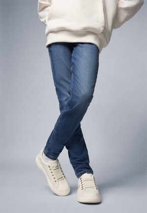 Blaue Denim-Jeans mit slim fit und einem ausgewaschenen Textur. Kombiniert mit weißen Sneakers, die goldene Akzente und ein gesticktes Design aufweisen.