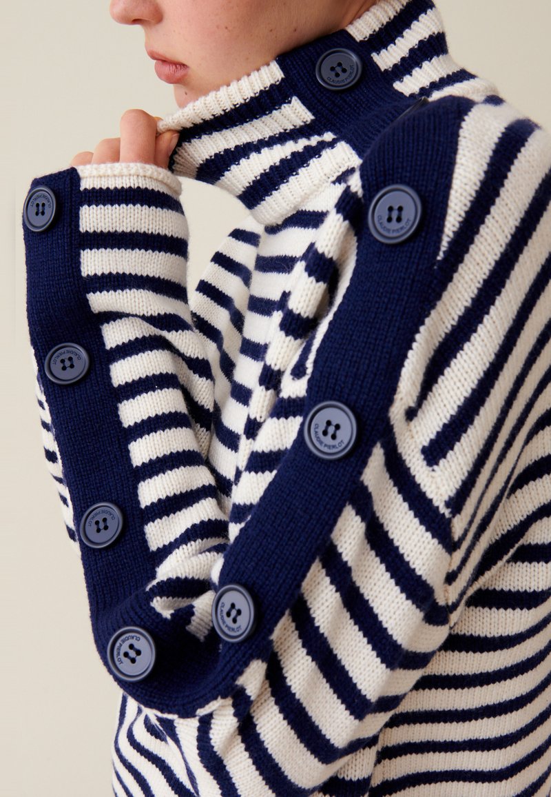 Marineblauer und weiß gestreifter Pullover mit hohem Kragen, großen Knöpfen an den Ärmeln und einer weichen Strickstruktur.