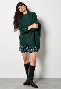 Maglione verde oversize a coste con collo alto, indossato sopra un vestito nero floreale con ruches, abbinato a stivali neri con tacco al ginocchio.