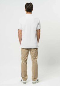 Mężczyzna stoi tyłem, ubrany w białą koszulkę polo, beżowe spodnie i białe trampki w niebieskie i czerwone paski.