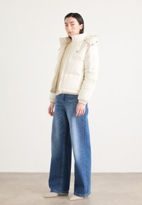 Polstret kremfarget jakke med hette, designet med glidelås foran og elastiske mansjetter, kombinert med vide blå denimbukser.