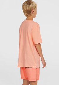 T-shirt a maniche corte di un leggero colore pesco con una vestibilità comoda, in cotone, design semplice, indossato con pantaloni a shorts pesco abbinati con un logo scuro.