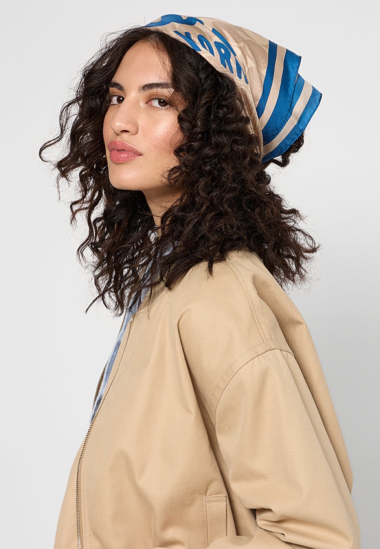 Veste bomber beige avec poches avant, associée à une écharpe à motifs beige et bleue comportant des lettres en gras. Des cheveux bouclés encadrent le visage.