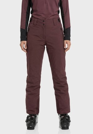 Person trägt dunkelweinrote Skihose und passende Jacke mit schwarzen Skischuhen und steht vor hellem Hintergrund.
