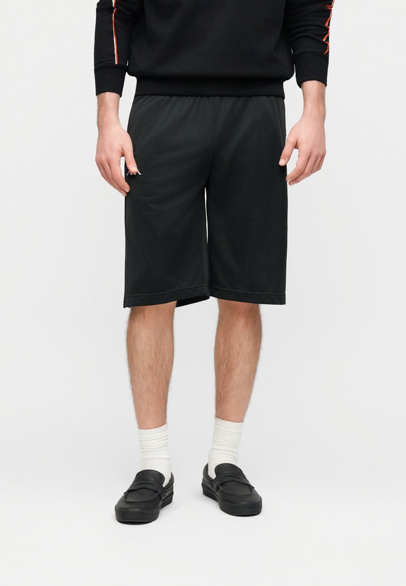 Άνδρας που φοράει μαύρο σορτς μέχρι το γόνατο, λευκές κάλτσες, μαύρα παπούτσια χωρίς κορδόνια, και μαύρο φούτερ με πορτοκαλί ρίγες στα μανίκια.