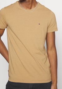 T-shirt en coton beige avec un col rond, des manches courtes et un petit logo multicolore sur la poitrine. Texture lisse, coupe ajustée, style décontracté.