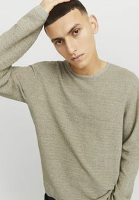 Hellbraun strukturierter Pullover mit rundem Halsausschnitt und langen Ärmeln. Das weiche Material hat dezente Muster. Das Modell posiert mit der Hand am Kopf.