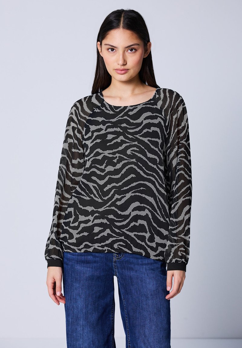 Top lungo trasparente nero e grigio con motivo a strisce zebra, scollatura tonda e vestibilità morbida, abbinato a pantaloni in denim blu.