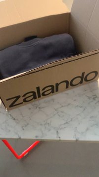Cardboard box with "zalando" printed in black, containing a folded dark gray sweatshirt, placed on a marble surface with red accents.