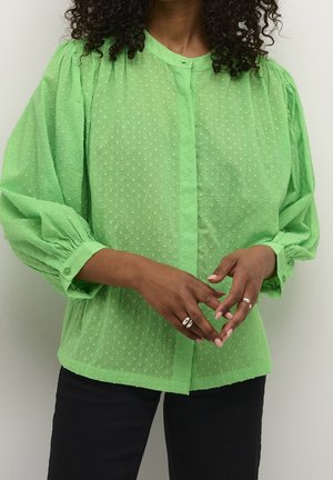 Blouse sheer verte à pois, avec des manches bouffantes et une fermeture à boutons, arborant une texture subtile et une coupe décontractée, associée à un pantalon noir.