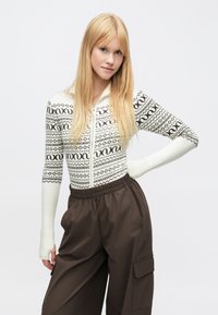 Džemperis ar rāvējslēdzēju un ģeometriskiem rakstiem krēmkrāsā un tumši brūnā krāsā, kopā ar brūnām cargo biksēm ar elastīgu vidukli un sānu kabatām.