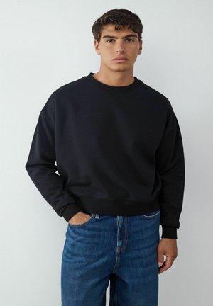 Schwarzer Rundhalsausschnitt-Sweatshirt mit langen Ärmeln und gerippten Bündchen, kombiniert mit Jeans. Der Stoff wirkt glatt und leicht tailliert.