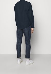 Námořnická košile na knoflíky s jemným vertikálním pruhovaným vzorem, v kombinaci s ohrnutými tmavými džínami a bílými teniskami.