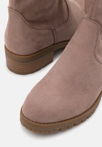 Botines de ante rosa claro con acabado texturizado, suelas de goma y un tacón ligeramente apilado. Presentan un detalle sencillo de costuras a lo largo de las costuras.