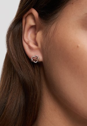 Nahaufnahme eines Frauenohrs mit einem kleinen herzförmigen Ohrstecker, verziert mit klaren Kristallen.