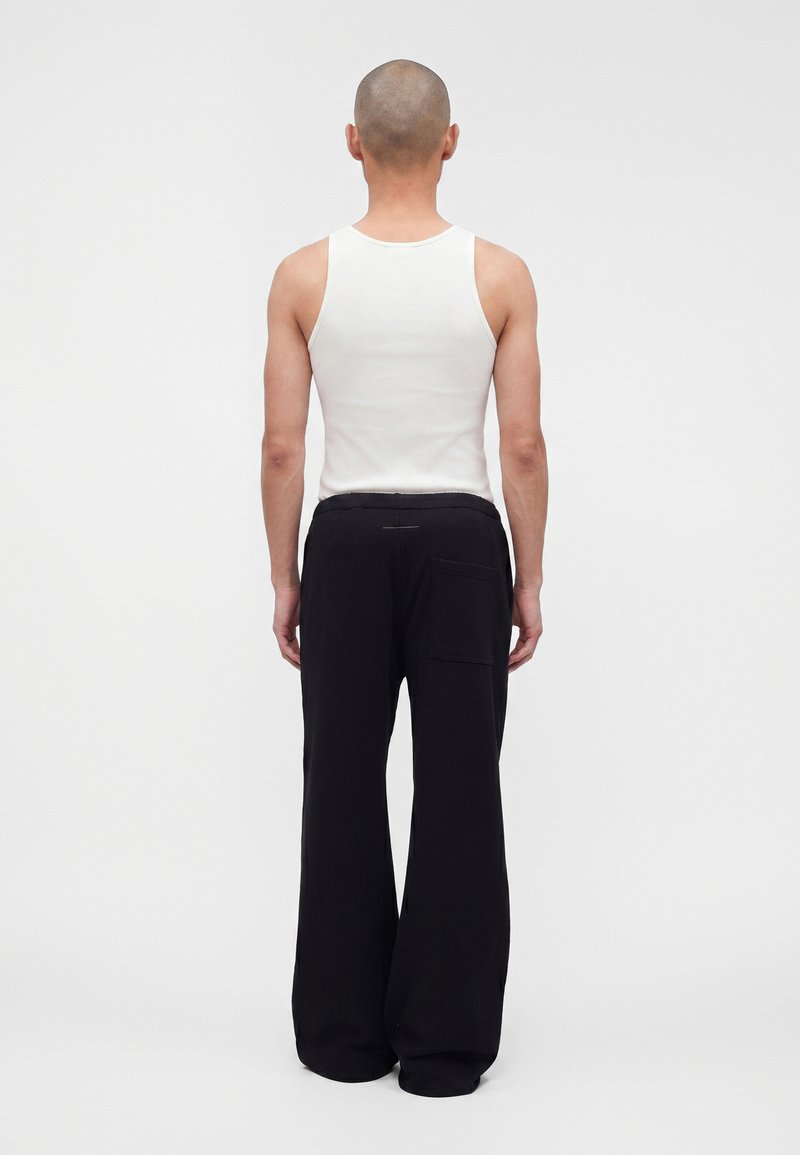 Personne à la tête rasée, vue de dos, portant un haut blanc sans manches et un pantalon noir ample avec une poche arrière, debout devant un fond uni.