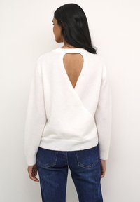 Hvid strikket sweater med en v-formet åben ryg. Ribbet kant og ærmer. Sammen med blå jeans, hvilket viser et afslappet look.