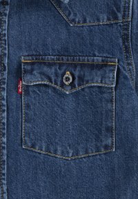Il tessuto in denim blu scuro presenta una tasca sul petto con un pattina appuntita, un bottone a pressione sicuro e cuciture gialle a contrasto. Etichetta Levi's visibile.