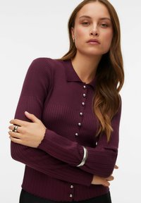 Bordo spalvos kotuotas megztinis su apykakle ir sidabro spalvos sagų užsegimais. Ilgos rankovės su banguotais kraštais. Lygus tekstūra.